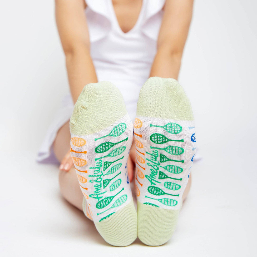 Child wearing white ankle socks with pastel green cuffs, showing colorful tennis racket patterns and “Ame & Lulu” text on the soles while sitting with feet pointed toward the camera.