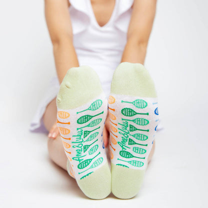 Child wearing white ankle socks with pastel green cuffs, showing colorful tennis racket patterns and “Ame & Lulu” text on the soles while sitting with feet pointed toward the camera.