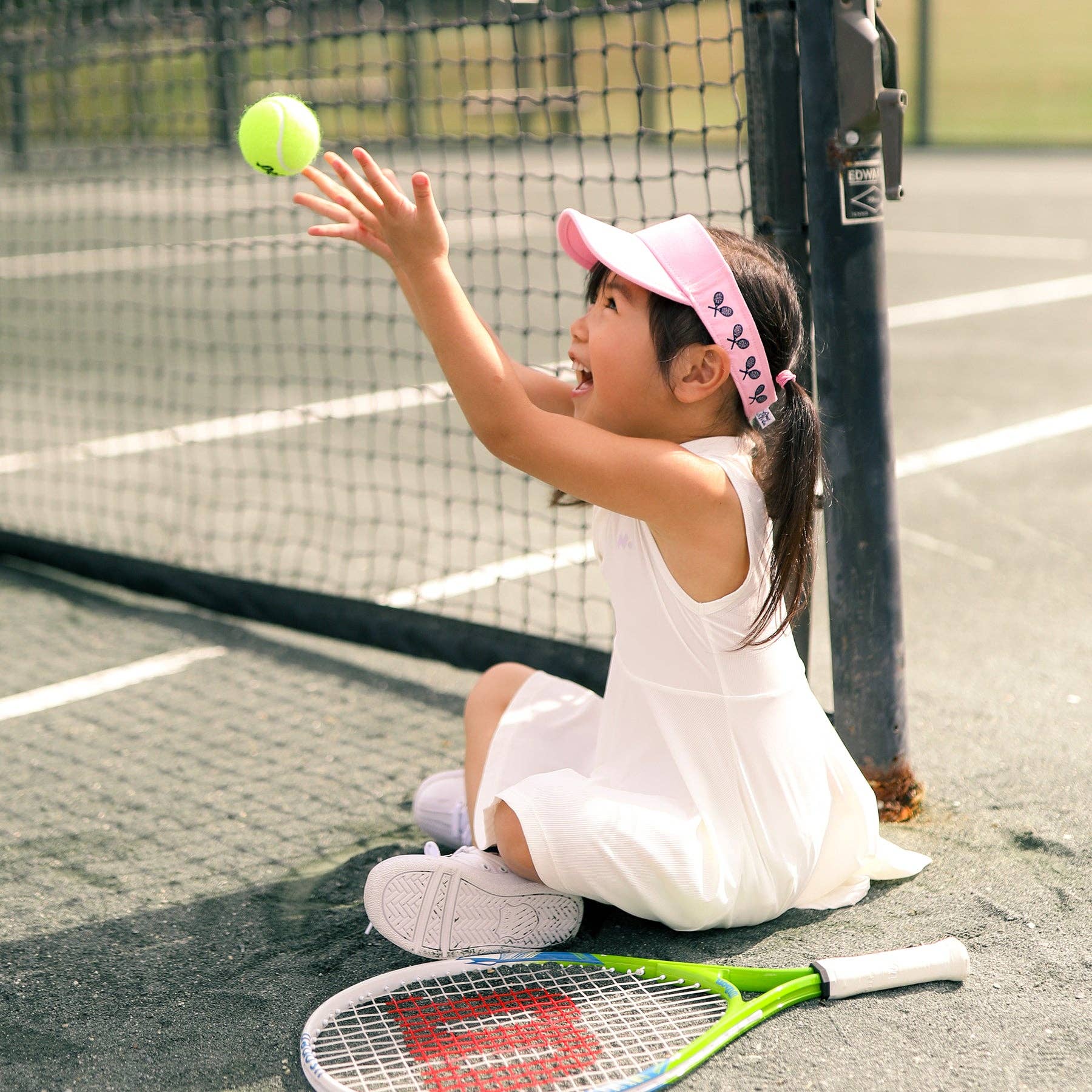 Girl wearing pink kids visor sitting on a tennis court tossing a tennis ball in the air