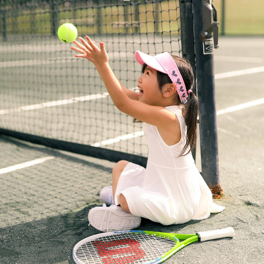 Girl wearing pink kids visor sitting on a tennis court tossing a tennis ball in the air