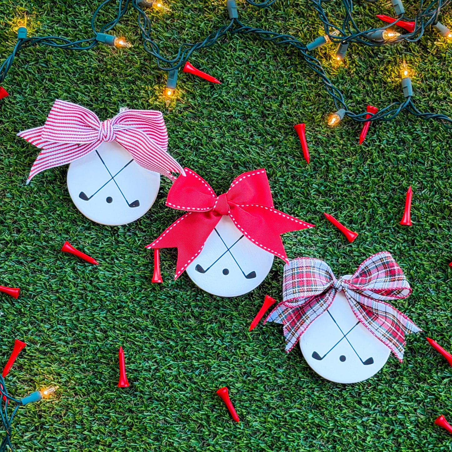 Three white ceramic ornaments featuring black crossed golf clubs, each with a different ribbon — red-and-white stripe, solid red, and tartan — arranged on green turf with holiday lights and scattered red golf tees.