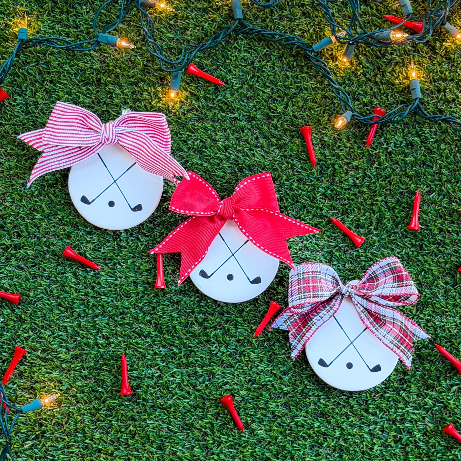 Three white ceramic ornaments featuring black crossed golf clubs, each with a different ribbon — red-and-white stripe, solid red, and tartan — arranged on green turf with holiday lights and scattered red golf tees.