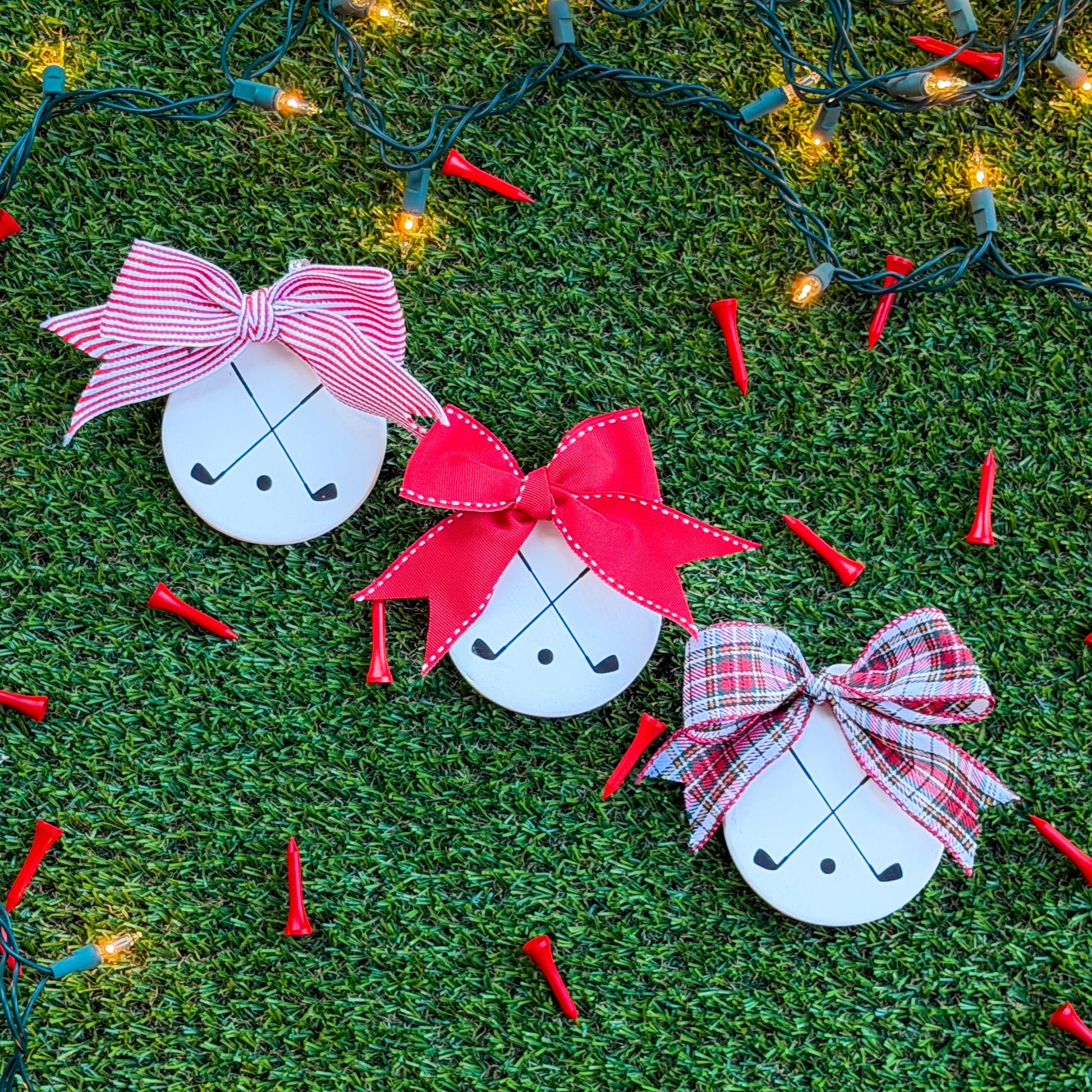 Three white ceramic ornaments featuring black crossed golf clubs, each with a different ribbon — red-and-white stripe, solid red, and tartan — arranged on green turf with holiday lights and scattered red golf tees.